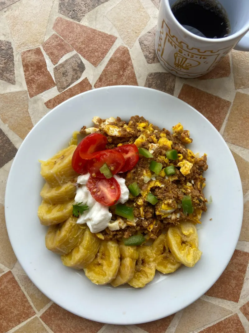 Prato com banana-da-terra, ovos mexidos com carne e tomate, ao lado de um café